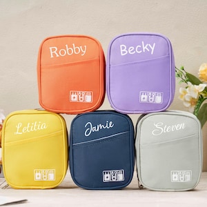 May include: A stack of six colorful travel pouches, each with a name printed on the front: Robby, Becky, Letitia, Jamie, and Steven. The pouches are orange, purple, yellow, navy blue, and gray. Each pouch has a small pocket and a white icon.