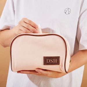 May include: A pink canvas makeup bag with a brown leather trim and a brown leather patch with the initials "DSH" embroidered on it.