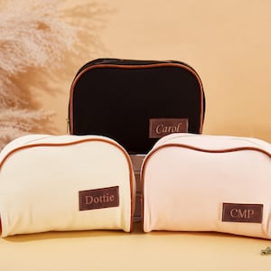 Könnte beinhalten: Drei Kosmetiktaschen aus Canvas mit braunem Lederbesatz und personalisierten Namen. Die Taschen sind in Schwarz, Creme und Rosa.