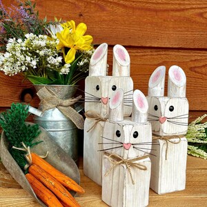 May include: Wooden bunny figures with pink-lined ears, black eyes, and pink noses. The bunnies have twine whiskers and are tied with twine bows. A metal scoop holds wooden carrots. A galvanized metal vase holds flowers.