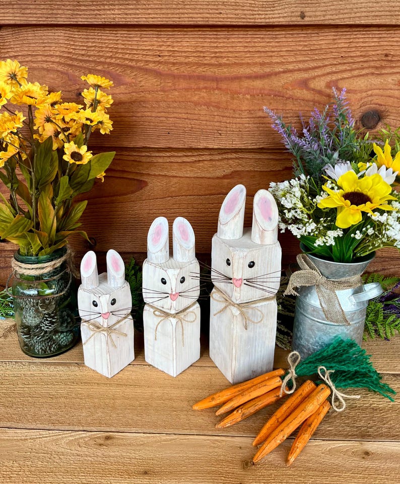 May include: Three white wooden bunny figures of varying sizes, with pink-lined ears, whiskers, and twine bows. Orange carrot decorations and floral arrangements in glass and metal containers complete the Easter-themed display.