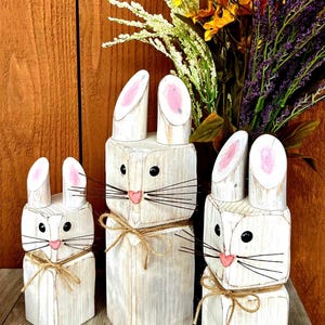 May include: Three white wooden bunny figures with pink inner ears, black eyes, and pink noses. Each bunny has a twine bow around its neck. The bunnies are of varying sizes and are displayed on a wooden surface.