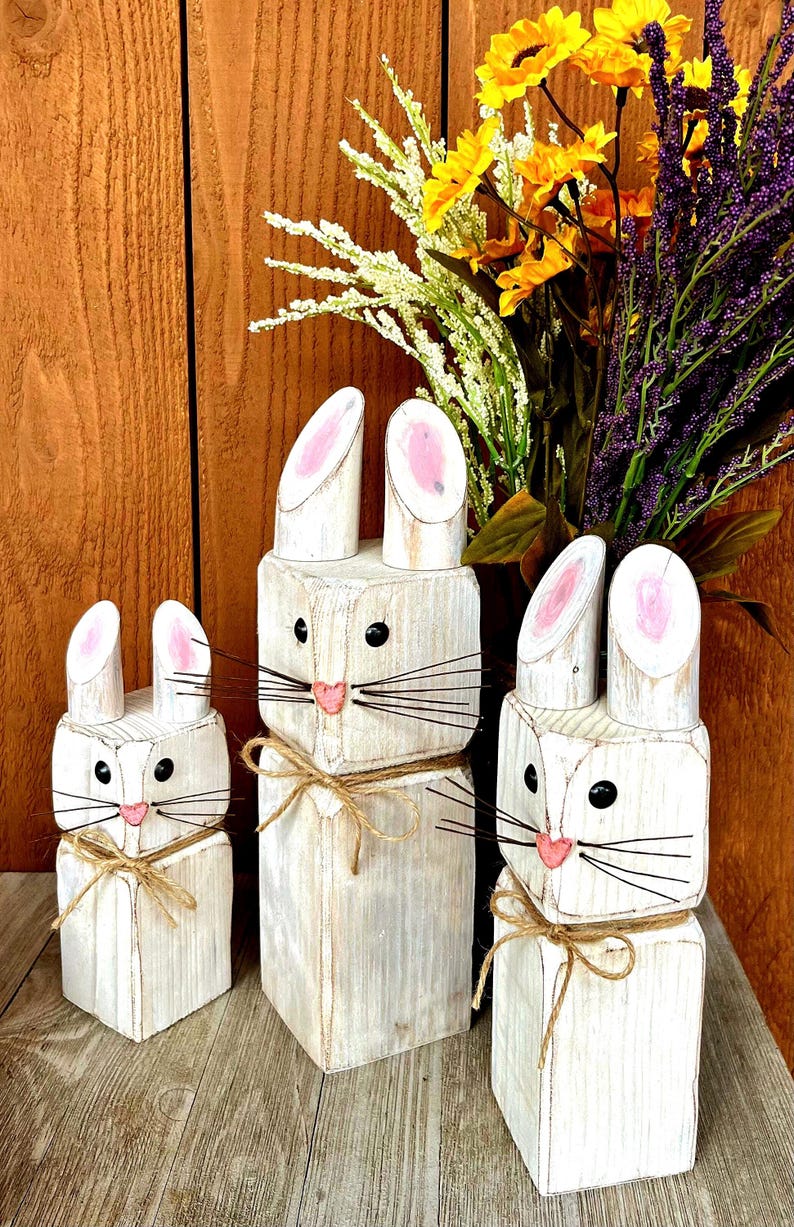 May include: Three white wooden bunny figures with pink inner ears, black eyes, and pink noses. Each bunny has a twine bow around its neck. The bunnies are of varying sizes and are displayed on a wooden surface.