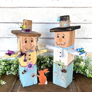 May include: Two wooden scarecrows with painted faces, straw hair, and hats. One wears a plaid scarf and a yellow shirt, the other a blue gingham scarf and a white shirt. A wooden orange bunny stands between them. Decorative butterflies and flowers are attached.