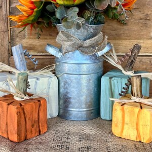 May include: Wooden pumpkin decorations in orange, yellow, and light blue, with straw and twig accents. A galvanized metal milk jug with a burlap bow and a floral arrangement complete the fall-themed display.