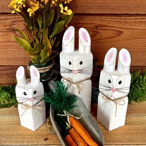 May include: Three white wooden bunny figures with pink-lined ears, black eyes, and whiskers. They are tied with twine bows. A metal scoop holds orange carrots and green foliage. Yellow flowers in a vase are in the background.