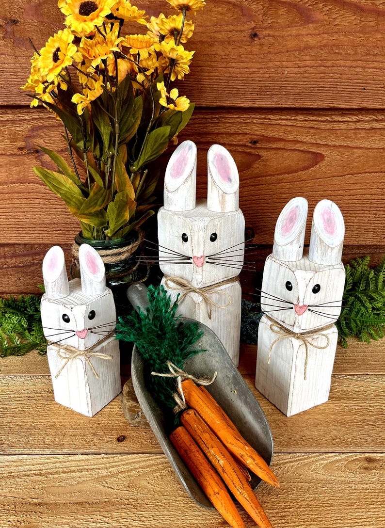 May include: Three white wooden bunny figures with pink-lined ears, black eyes, and whiskers. They are tied with twine bows. A metal scoop holds orange carrots and green foliage. Yellow flowers in a vase are in the background.