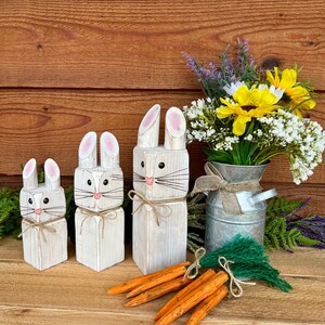 May include: Three white wooden bunny figures with pink inner ears and whiskers, arranged on a wooden surface. Orange wooden carrots and a bouquet of flowers in a metal container complete the Easter-themed display.