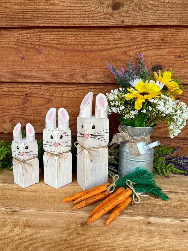 May include: Three white wooden bunny figures with pink inner ears and whiskers, arranged on a wooden surface. Orange wooden carrots and a bouquet of flowers in a metal container complete the Easter-themed display.