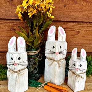 May include: Three white wooden bunny figures with pink inner ears, black eyes, and twine whiskers and bows. Orange wooden carrots are in front of the bunnies. Yellow flowers in a green jar are in the background.