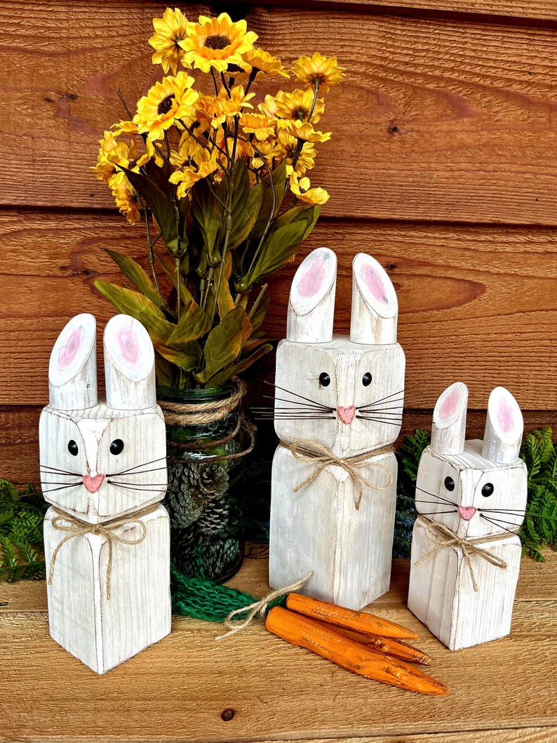 May include: Three white wooden bunny figures with pink inner ears, black eyes, and twine whiskers and bows. Orange wooden carrots are in front of the bunnies. Yellow flowers in a green jar are in the background.