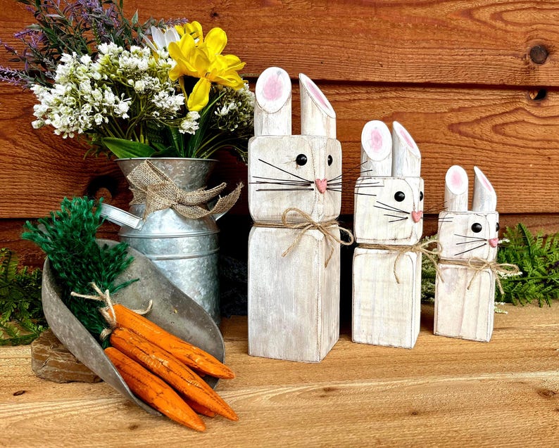 May include: Wooden Easter bunny figurines in varying sizes, painted white with pink accents on the ears and noses. Each bunny has black whiskers and is tied with twine. A metal scoop holds orange carrots, and a galvanized metal vase displays flowers.