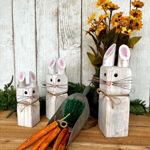 May include: Wooden bunny figurines with pink-lined ears, black whiskers, and twine accents. A metal scoop holds faux carrots and green filler. Yellow flowers in a vase complete the Easter-themed display.