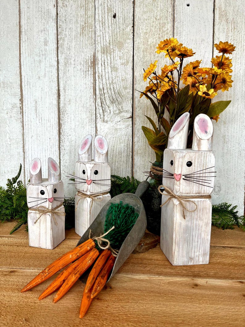 May include: Wooden bunny figurines with pink-lined ears, black whiskers, and twine accents. A metal scoop holds faux carrots and green filler. Yellow flowers in a vase complete the Easter-themed display.