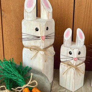 May include: Two white wooden bunny figures with pink inner ears, black eyes, and pink noses. The bunnies have wire whiskers and are tied with twine. A small metal tray holds wooden carrots and green yarn.