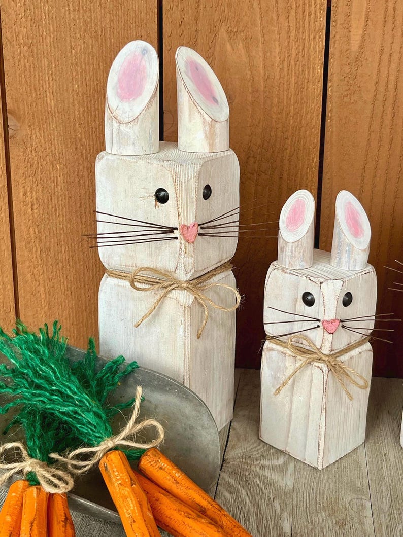 May include: Two white wooden bunny figures with pink inner ears, black eyes, and pink noses. The bunnies have wire whiskers and are tied with twine. A small metal tray holds wooden carrots and green yarn.
