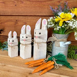 May include: Three wooden bunny figures with pink-lined ears, black eyes, and pink noses. Each bunny has a twine bow. Orange wooden carrots and a metal vase with flowers complete the Easter-themed display.