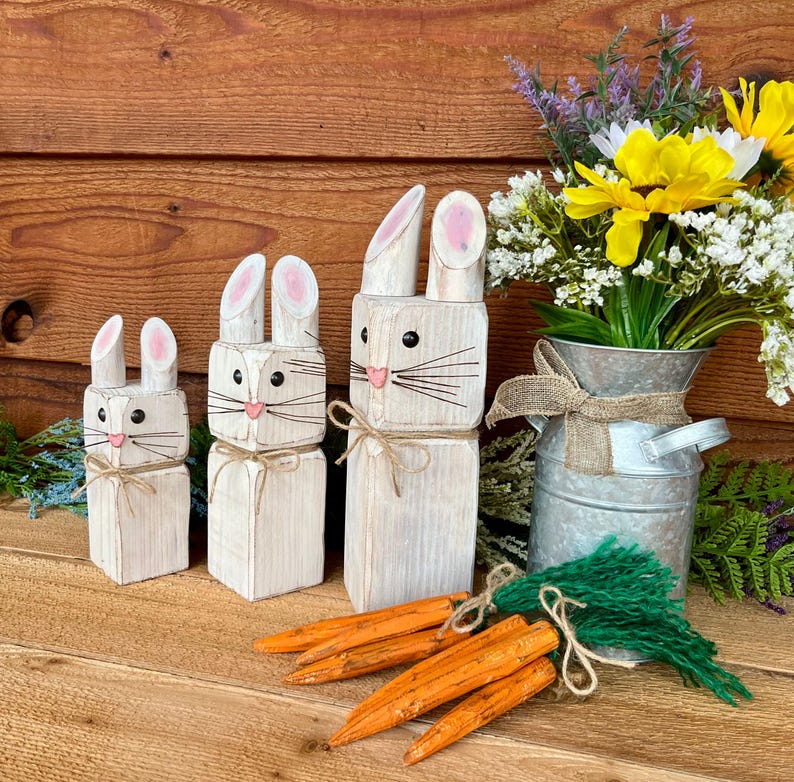 May include: Three wooden bunny figures with pink-lined ears, black eyes, and pink noses. Each bunny has a twine bow. Orange wooden carrots and a metal vase with flowers complete the Easter-themed display.