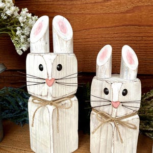 May include: Two white wooden bunny figures with black eyes, pink noses, and wire whiskers. The ears are made of two vertical pieces of wood with pink interiors. Each bunny has a twine bow around its neck. The bunnies are on a wooden surface.