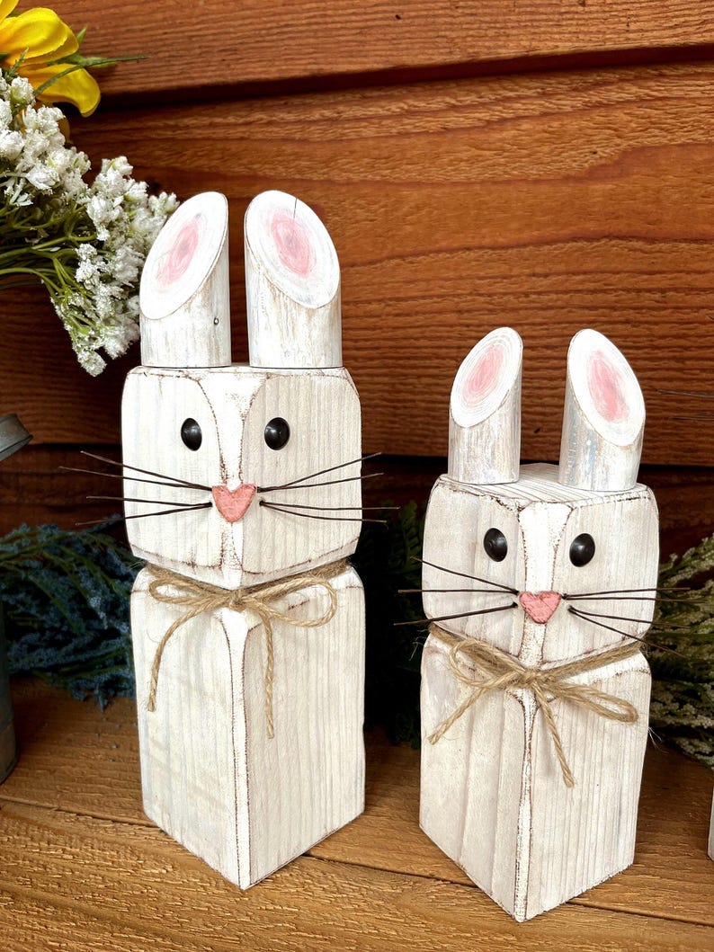 May include: Two white wooden bunny figures with black eyes, pink noses, and wire whiskers. The ears are made of two vertical pieces of wood with pink interiors. Each bunny has a twine bow around its neck. The bunnies are on a wooden surface.