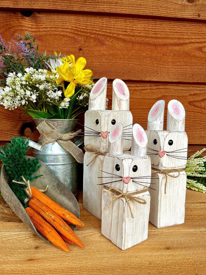 May include: Wooden bunny figures with pink-lined ears, black eyes, and pink noses. The bunnies have twine whiskers and are tied with twine bows. A metal scoop holds wooden carrots. A galvanized metal vase holds flowers.