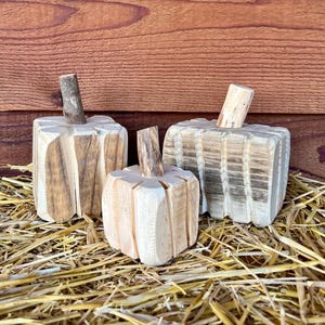 May include: Three carved wooden pumpkins with natural light wood grain and distinct vertical grooves. Each pumpkin features a small, cylindrical branch segment as a stem, showcasing varied wood tones and textures in a rustic, handcrafted style.