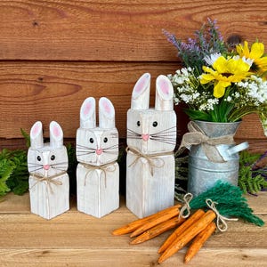 May include: Three white wooden bunny figures with pink-tipped ears and twine accents. The bunnies are arranged in a row, with a bouquet of flowers in a metal pitcher and wooden carrots.