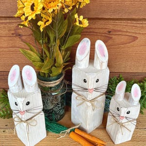 May include: Three white wooden bunny figures with pink inner ears and twine whiskers. Each bunny has a twine bow and is paired with orange wooden carrots. A bouquet of yellow sunflowers sits in a glass jar.