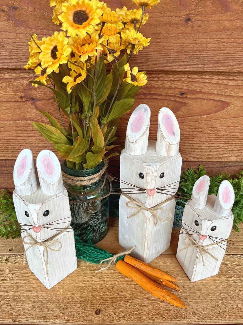 May include: Three white wooden bunny figures with pink inner ears and twine whiskers. Each bunny has a twine bow and is paired with orange wooden carrots. A bouquet of yellow sunflowers sits in a glass jar.