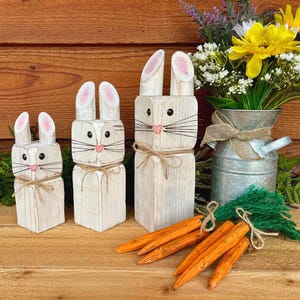 May include: Three wooden bunny figures with pink-tipped ears, black eyes, and pink noses. Each bunny is tied with twine. Orange wooden carrots and a bouquet of yellow flowers in a metal vase complete the Easter-themed display.