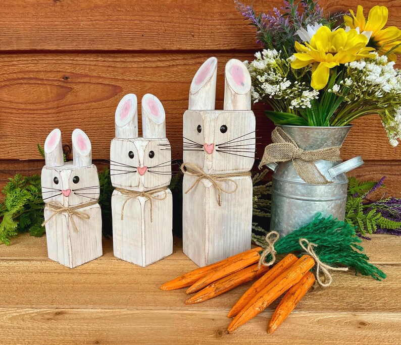May include: Three wooden bunny figures with pink-tipped ears, black eyes, and pink noses. Each bunny is tied with twine. Orange wooden carrots and a bouquet of yellow flowers in a metal vase complete the Easter-themed display.