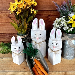 May include: Three white wooden bunny figures with pink-lined ears, black eyes, and whiskers, tied with twine. A metal scoop holds orange carrots with green tops. Yellow and white flowers in vases complete the Easter-themed display.