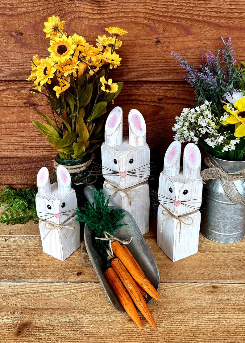 May include: Three white wooden bunny figures with pink-lined ears, black eyes, and whiskers, tied with twine. A metal scoop holds orange carrots with green tops. Yellow and white flowers in vases complete the Easter-themed display.