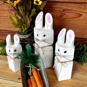 May include: Wooden bunny figurines with pink-painted inner ears, black whiskers, and twine bows. A metal scoop holds orange carrot decorations with green tops. Yellow flowers in a green vase are in the background. The bunnies are painted white.