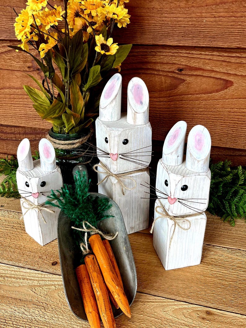 May include: Wooden bunny figurines with pink-painted inner ears, black whiskers, and twine bows. A metal scoop holds orange carrot decorations with green tops. Yellow flowers in a green vase are in the background. The bunnies are painted white.