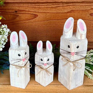 May include: Three wooden bunny figures in varying sizes. Each bunny is painted white with pink inner ears and a pink nose. They have black eyes and whiskers made of thin wire, tied with twine.