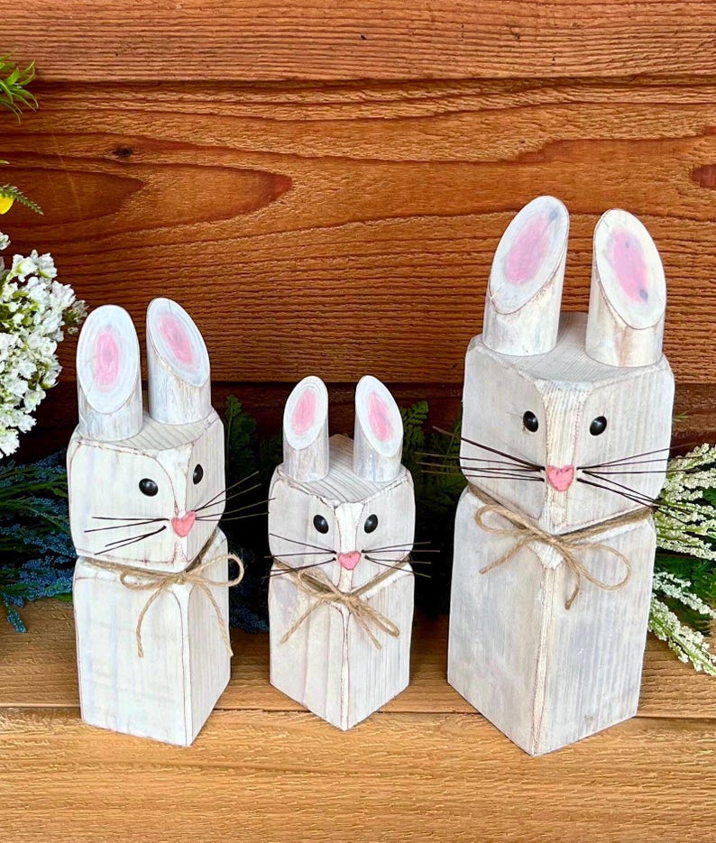 May include: Three wooden bunny figures in varying sizes. Each bunny is painted white with pink inner ears and a pink nose. They have black eyes and whiskers made of thin wire, tied with twine.