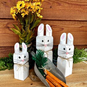 May include: Three white wooden bunny figures with pink-lined ears and black whiskers. A metal scoop holds orange carrots and green foliage. Yellow flowers in a vase are in the background. The bunnies are tied with twine.