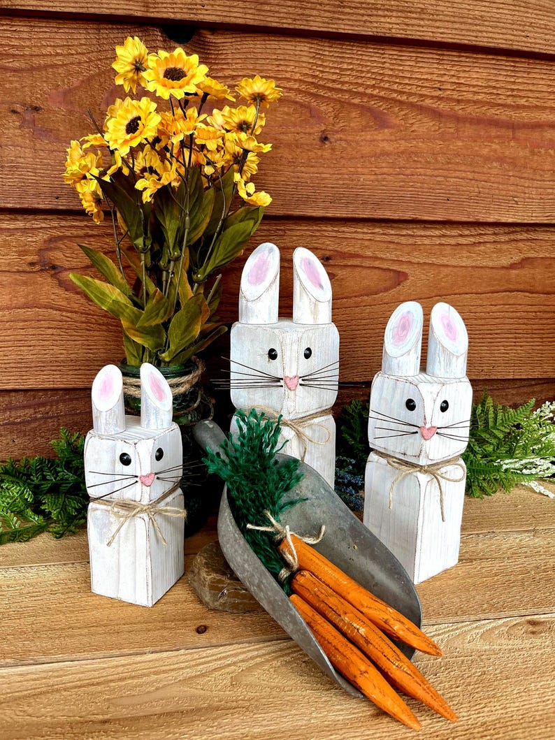 May include: Three white wooden bunny figures with pink-lined ears and black whiskers. A metal scoop holds orange carrots and green foliage. Yellow flowers in a vase are in the background. The bunnies are tied with twine.