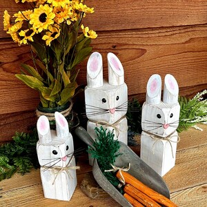 May include: Three white wooden bunny figures with pink inner ears, black eyes, and twine accents. A metal scoop holds orange carrots and green foliage. Yellow sunflowers in a green vase are in the background. The items are on a wooden surface.