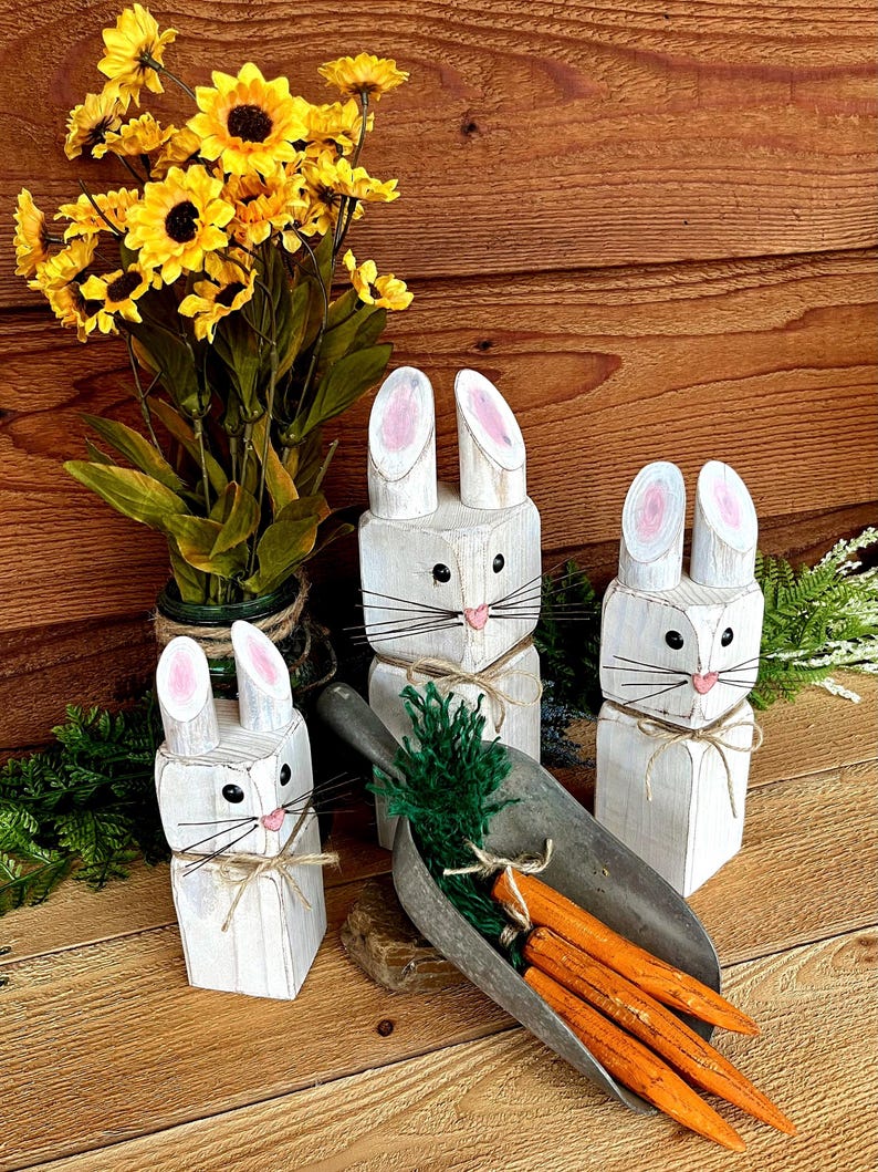 May include: Three white wooden bunny figures with pink inner ears, black eyes, and twine accents. A metal scoop holds orange carrots and green foliage. Yellow sunflowers in a green vase are in the background. The items are on a wooden surface.