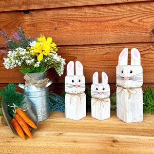 May include: Three white wooden bunny figures with pink inner ears and whiskers, tied with twine, stand next to a metal pail with flowers and a scoop of orange carrots. The bunnies are of varying sizes, perfect for Easter decor.