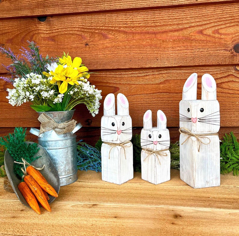 May include: Three white wooden bunny figures with pink inner ears and whiskers, tied with twine, stand next to a metal pail with flowers and a scoop of orange carrots. The bunnies are of varying sizes, perfect for Easter decor.