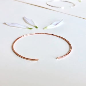 May include: A simple, hammered rose gold bangle bracelet. The bracelet is made of a thin, delicate wire and has a slightly rounded shape.