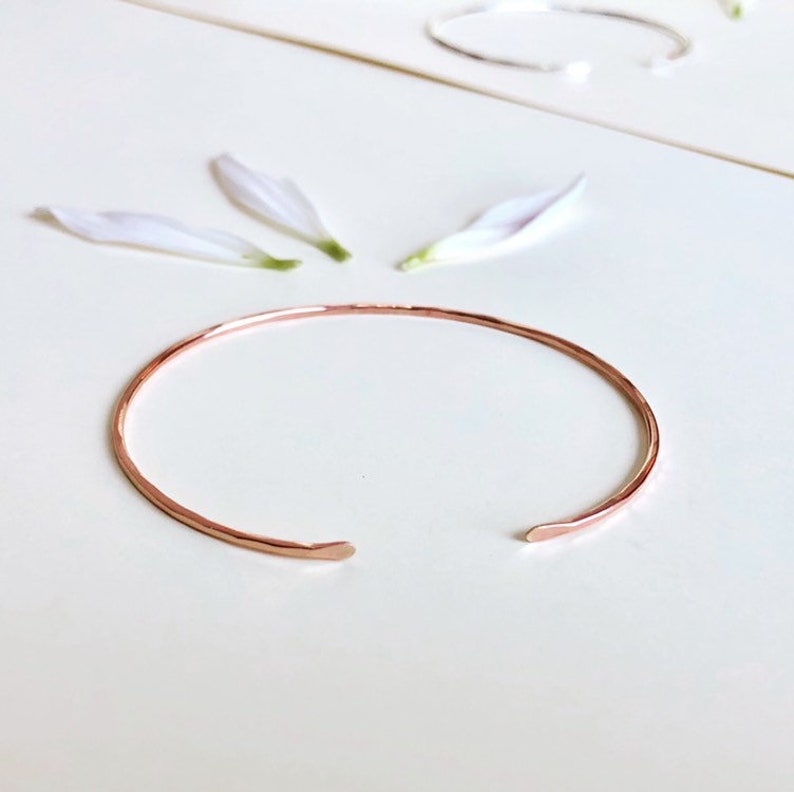 May include: A simple, hammered rose gold bangle bracelet. The bracelet is made of a thin, delicate wire and has a slightly rounded shape.