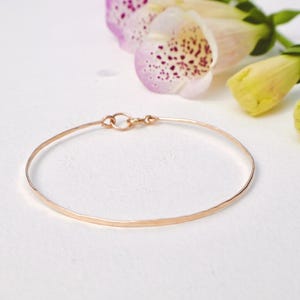 May include: A delicate, thin gold-colored bracelet with a simple clasp. The bracelet is set against a white background, with soft focus flowers in the background. The bracelet is a minimalist piece of jewelry.