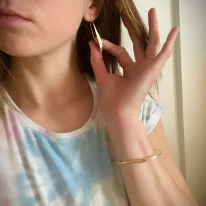 K&ouml;nnte beinhalten: Goldfarbene, l&auml;ngliche Ohrringe in Tropfenform. Ein goldenes Armband ist ebenfalls sichtbar. Die Person tr&auml;gt ein Tanktop mit Batikmuster. Die Ohrringe werden von einer Hand gehalten.