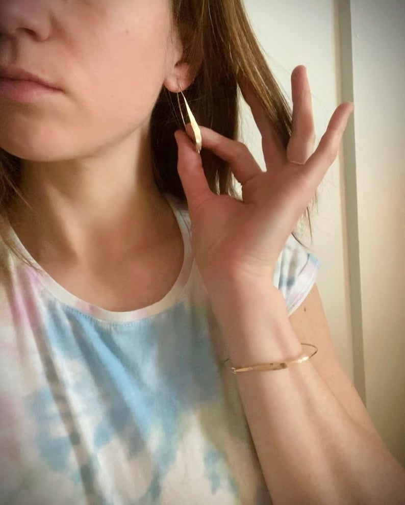 K&ouml;nnte beinhalten: Goldfarbene, l&auml;ngliche Ohrringe in Tropfenform. Ein goldenes Armband ist ebenfalls sichtbar. Die Person tr&auml;gt ein Tanktop mit Batikmuster. Die Ohrringe werden von einer Hand gehalten.