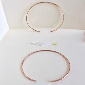 May include: Two gold-toned metal bangles, one thicker than the other, are displayed on a white surface. The bangles are open and circular in shape.