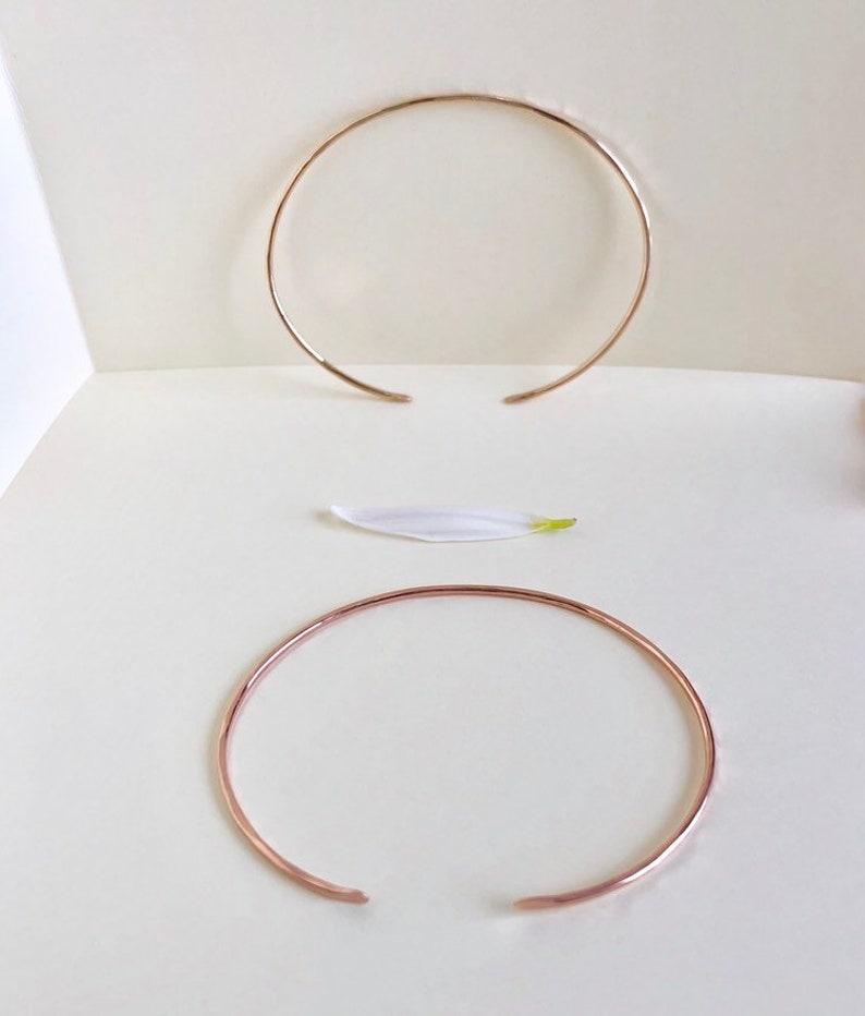 May include: Two gold-toned metal bangles, one thicker than the other, are displayed on a white surface. The bangles are open and circular in shape.